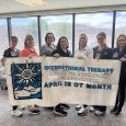 Seven ladies stand behind a banner celebrating Occupational Therapy month.