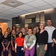 Sixteen employees together in front of hospital logo in main lobby.